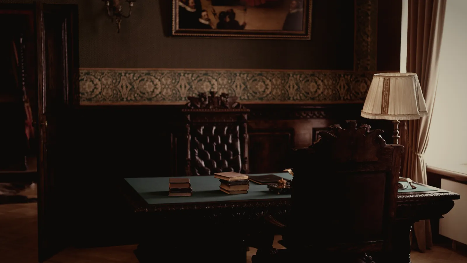A wood-paneled study with a carved desk, stacked books and a lamp