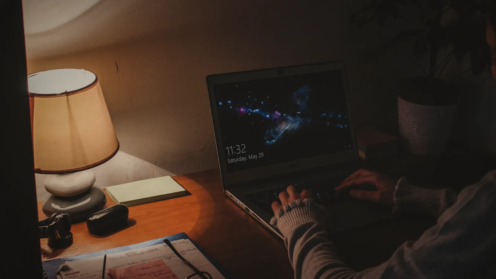 Hands typing on a laptop beside a warm table lamp in a home office at night