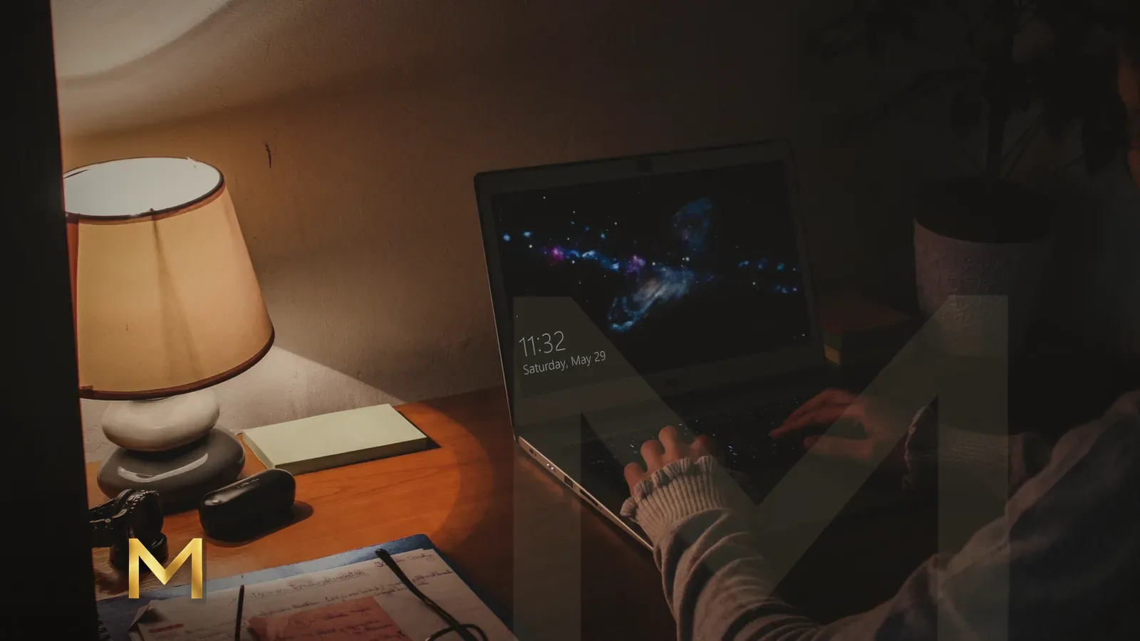 Hands typing on a laptop beside a warm table lamp in a home office at night