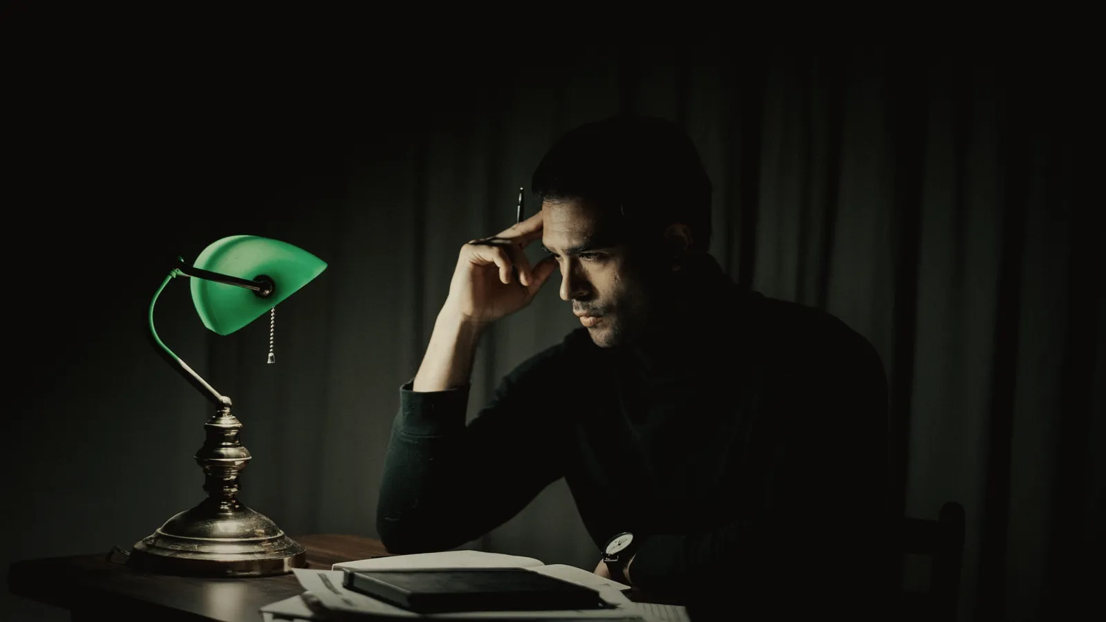 A man working alone at a desk under a banker's lamp late at night