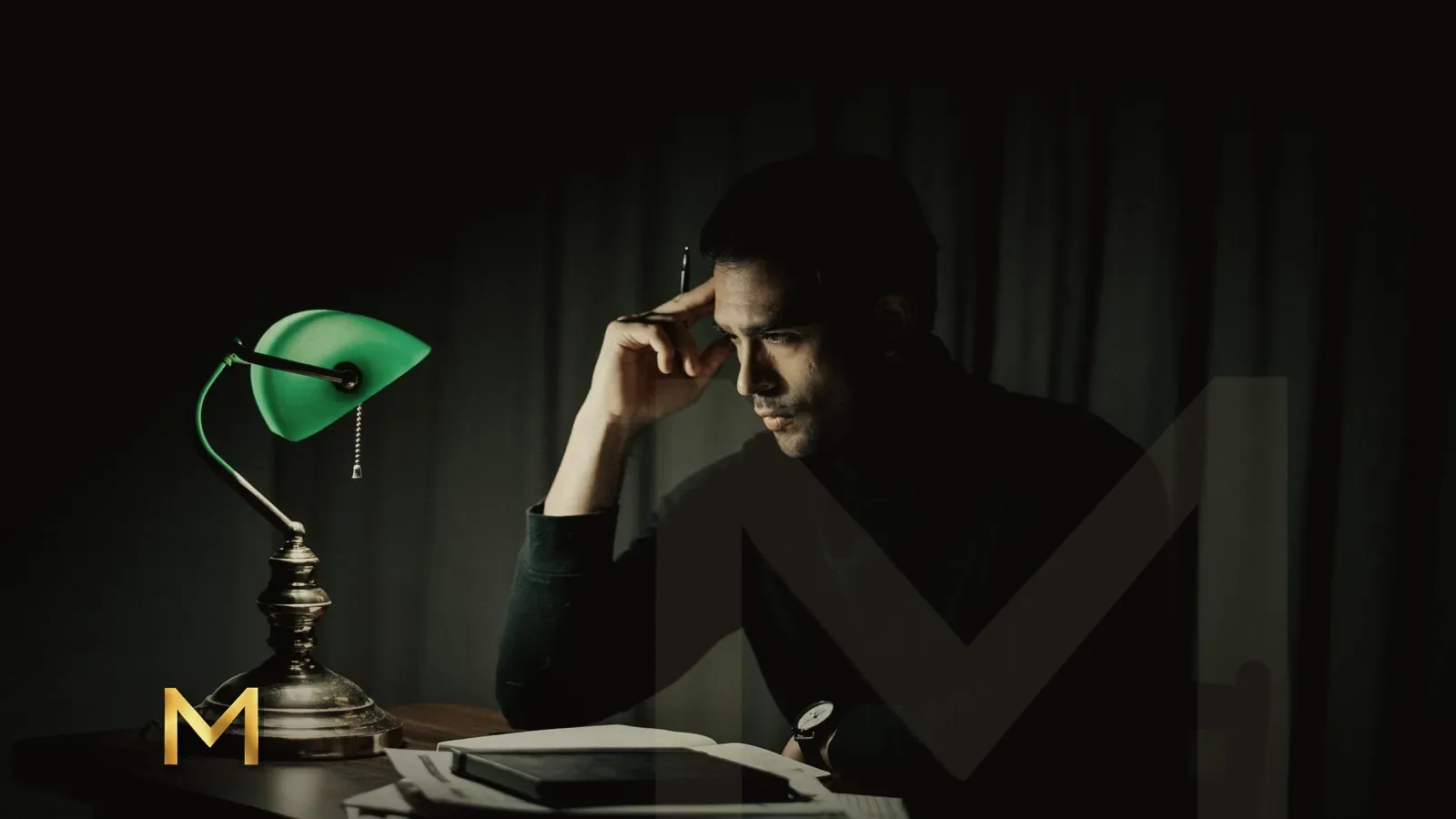 A man working alone at a desk under a banker's lamp late at night
